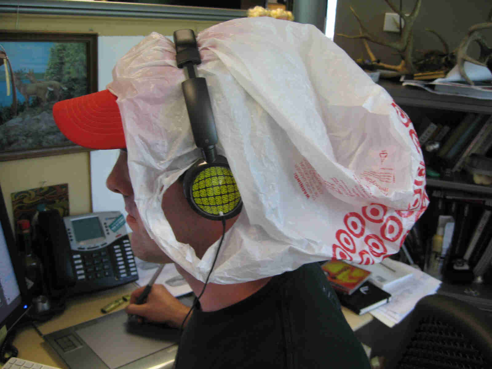 Left headshot of a person sitting at an office desk, wearing a plastic shopping bag on their head and headphones